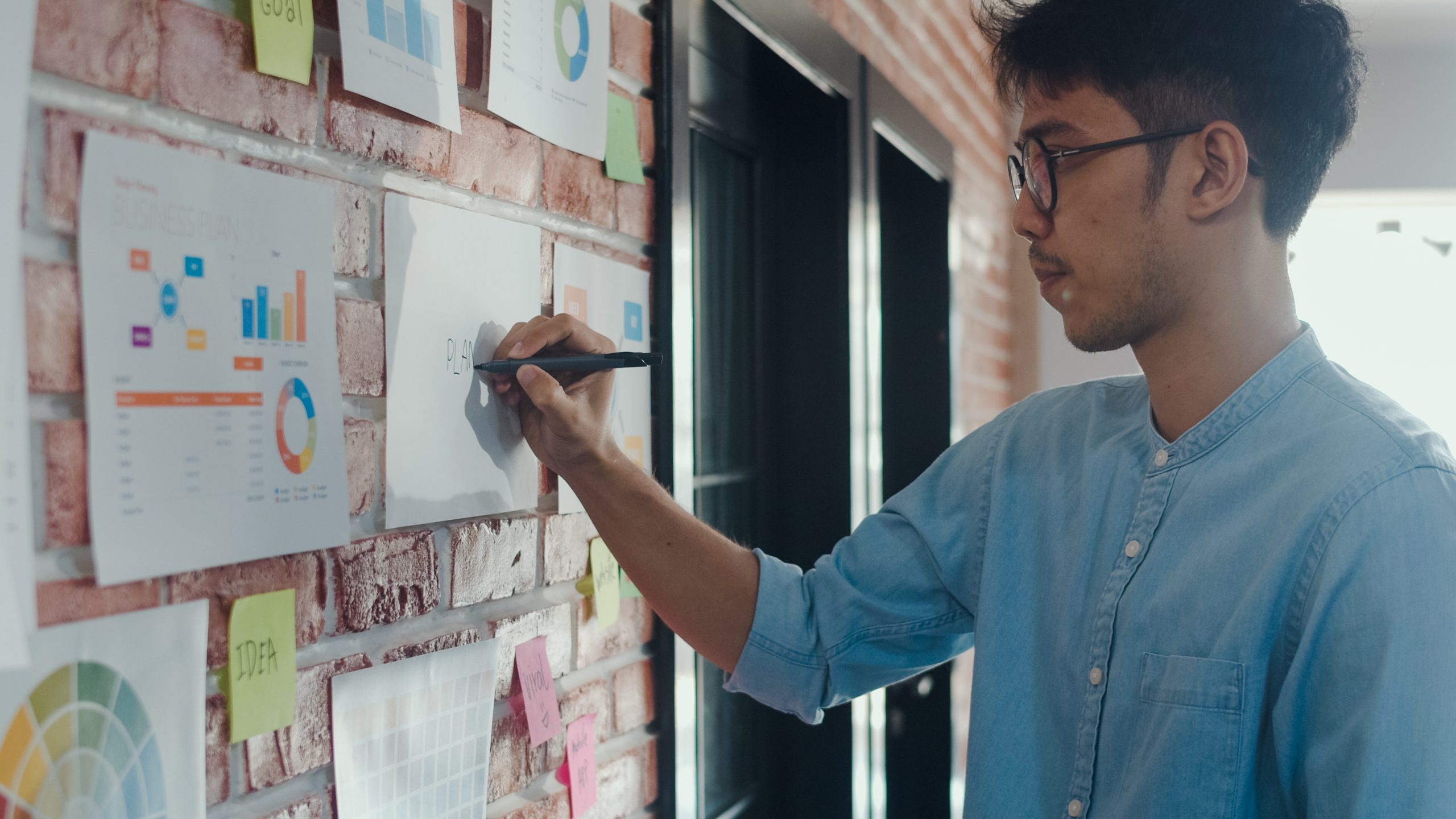 Asian creative man drawing work plan on paper board. Young professional business male think and write information reminder on paper on brick, business situation, startup in Loft office concept.