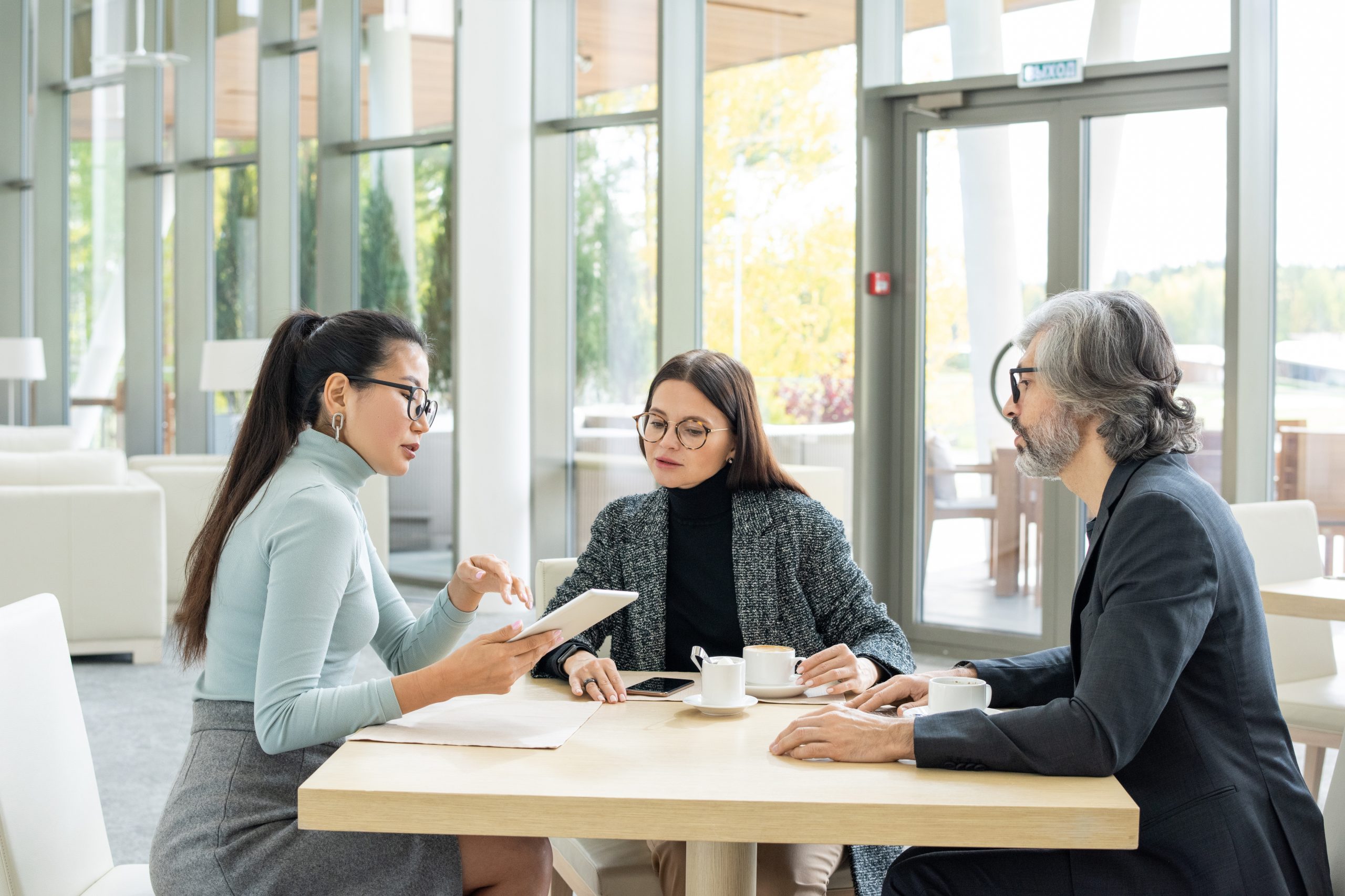 Confident Asian businesswoman in casualwear holding touchpad while sitting by table in front of two colleagues and presenting new plan